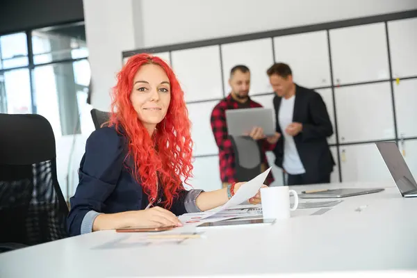 Red-haired woman boss sitting at working place with many papers and looking at camera with smile and confidence, glad to work with high-qualified professionals