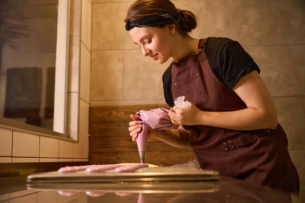 Pleased female baker squeezing macaron batter from pastry bag onto baking mat