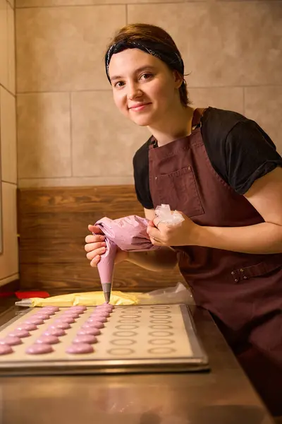 Female baker smiling at camera while squeezing macaron batter from piping bag onto baking mat