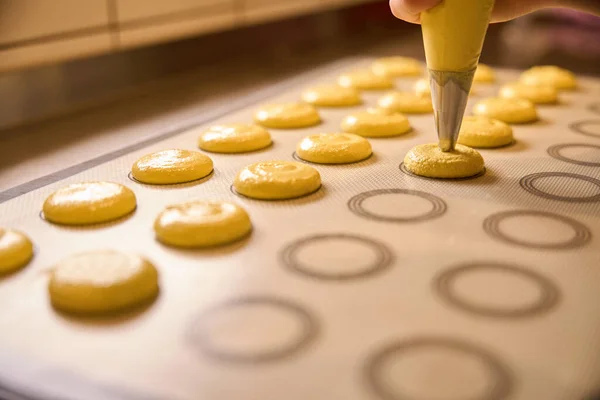 Closeup of hand squeezing yellow macaron batter from pastry bag onto silicone baking mat
