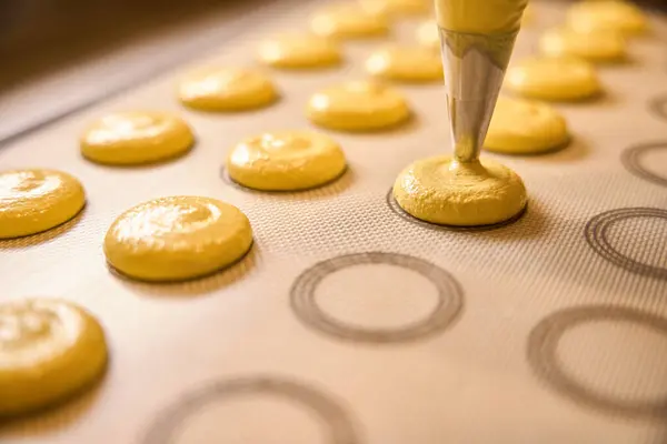 Closeup of lemon macaron batter being squeezed through pastry bag tip onto silicone baking mat