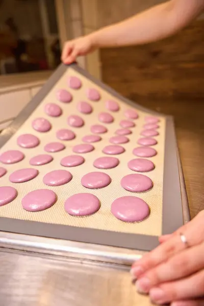 Closeup of pastry chef hands removing silicone mat with pink macaron shells from kitchen countertop