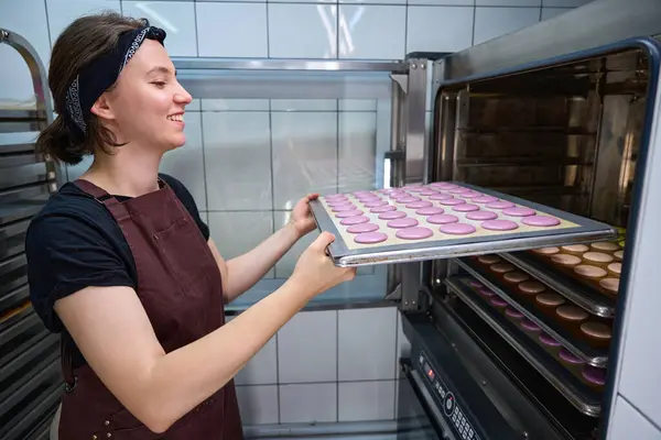 Side view of smiling pastry chef placing sheet pan with with macaron shells into combi-steam oven