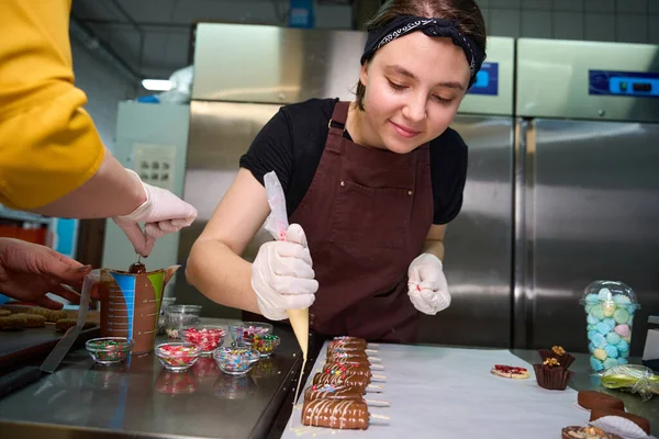 Pastry cook and her colleague in disposable gloves coating cakesicles with frosting and sprinkles
