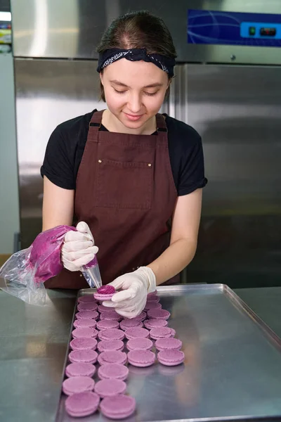 Pastry cook in disposable gloves piping purple filling on macaron shell over sheet pan on kitchen countertop