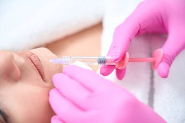 Kanepede yatan bir kadın, kozmetikçi elinde şırıngayla dudaklarına iğne yapıyor. Dudak büyütme