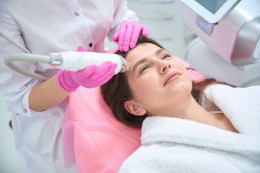 Kanepede uzanmış bir kadın portresi, güzellik uzmanı kucağında bir cihaz ve müşteriye yüz gerdirme hareketi.