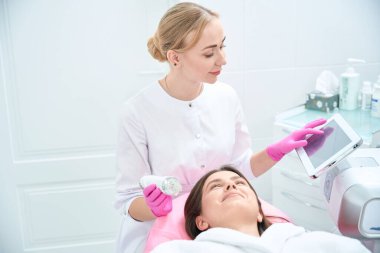Beautician holding aygıtı, istemcinin yanında oturuyor ve parametreleri ekrandan ayarlıyor