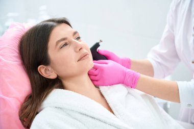 Kanepede uzanmış bir kadın, güzellik aletini tutuyor ve güzellik salonundaki müşteriye kulak deldirici yapıyor.