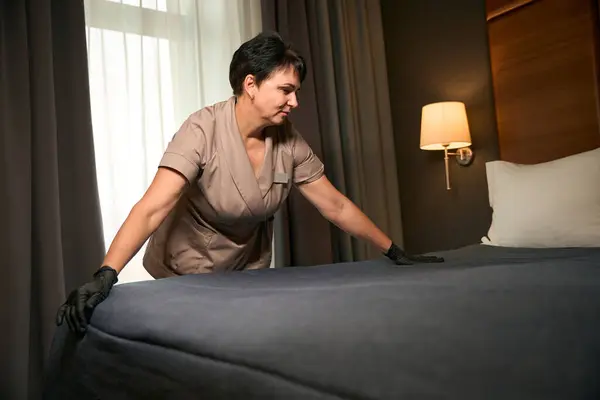 Focused chambermaid in disposable gloves and uniform making bed with bedspread in suite