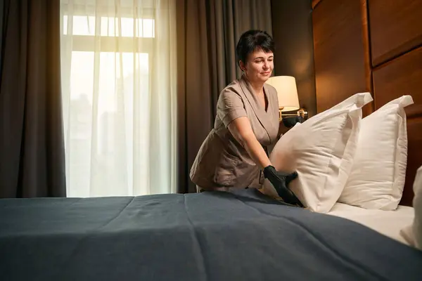 Pleased chambermaid in uniform and disposable gloves arranging pillows on bed in suite