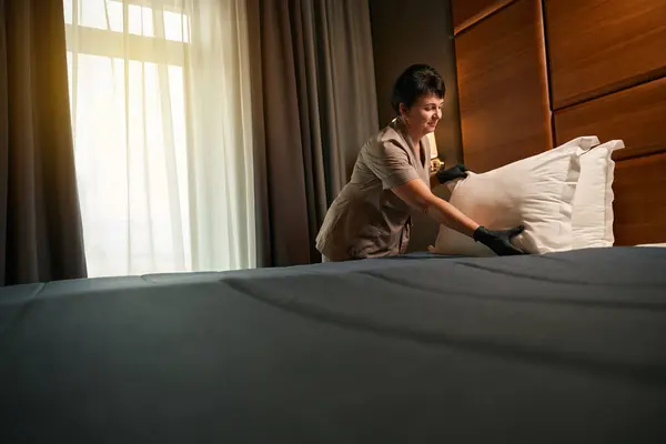 Contented maid in uniform and disposable gloves arranging pillows on hotel bed