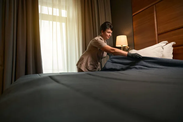 Chambermaid wearing uniform and disposable gloves while making hotel bed with bedcover