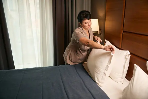 Tranquil chambermaid dressed in uniform placing pillows at head of bed in hotel room