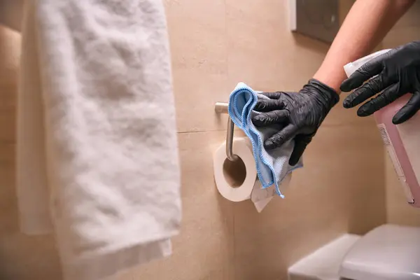 Cropped photo of gloved hands wiping wall-mounted toilet paper holder with microfiber cloth