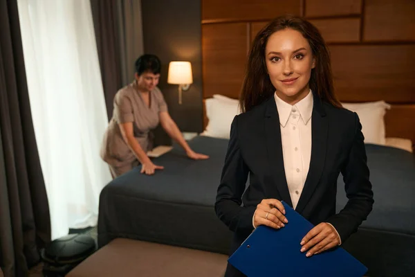 Pleased hotel manager with clipboard in hands standing in guest suite while chambermaid making bed