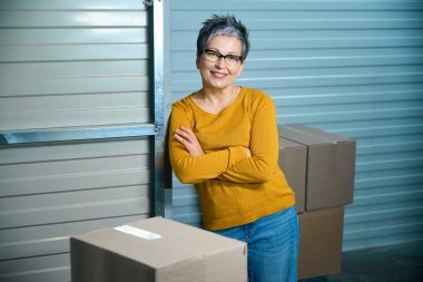 Karton bir kutunun yanındaki depoda duvara yaslanmış bir kadın.