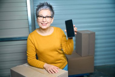 Depoda elinde telefonla kameraya bakan bir kadın.