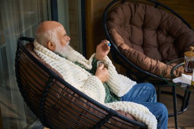 Verandada soğuk çay içen emekli adamın elinde bir termometre var.