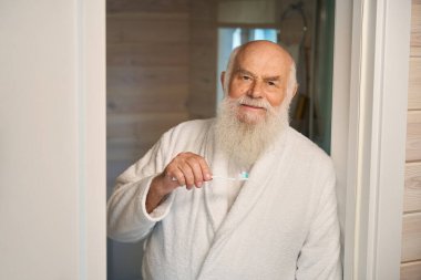 Gri sakallı yaşlı adam sabah rutinini banyoda yapıyor. Elinde diş fırçası var.