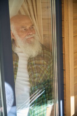 Gri saçlı, kareli gömlekli yaşlı adam pencereden dışarı bakar, modern ahşap bir evde yaşar.