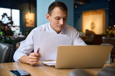 Mutfak çalışanı belgelerle çalışıyor. Lokanta salonunda dizüstü bilgisayarı var.