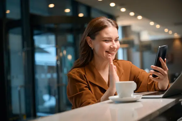 Joyful woman sitting at counter in hotel restaurant and waving her hand to conversation partner during videocall