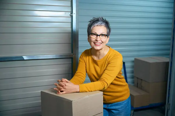 Woman with a short haircut and glasses looks smiling at the camera while leaning on boxes with things.