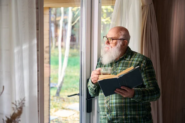 Pensioner enjoys a cup of tea by the window, he has a book in his hand