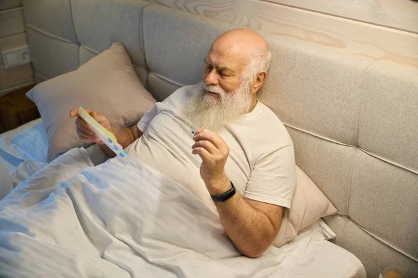 Old man holds a tablet box and a pill in his hands, he is located on a large bed
