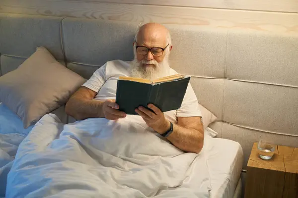 Old man reads a book before going to bed, he lies in a comfortable bed