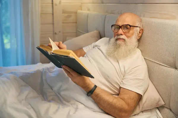 Gray-bearded old man with glasses reading a book before going to bed, he lies in a comfortable bed at home