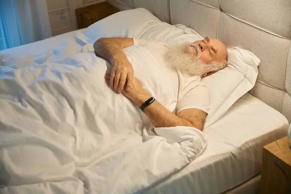 Old man sleeps on his back in comfortable bed, with a glass of water on the nightstand next to him