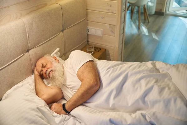 Gray-bearded old man sleeps in a comfortable bed, with a glass of water on the nightstand next to him