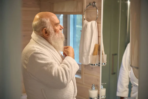 Old man in a white coat in the bathroom in front of the mirror combs his beard, he is in a white bathrobe