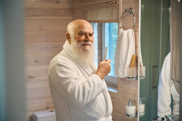Happy old man in bathroom stands in front of mirror with a comb in his hands, he is in a white bathrobe