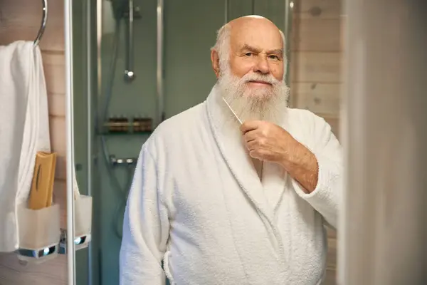 Gray-haired old man combs his beard in front of a mirror, he is in a white bathrobe
