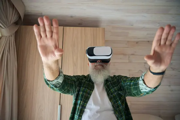 Gray-bearded old man in virtual reality glasses, he plays in a cozy bright room