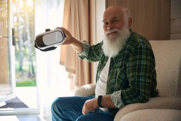 Happy gray-bearded old man holds virtual reality glasses in his hand, he sits in a cozy bright room