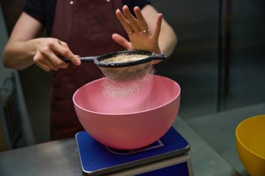 Önlüklü bir pasta şefinin mutfak terazisine koyduğu kase üzerinde elek ile badem unu ellerken çekilmiş fotoğrafı.