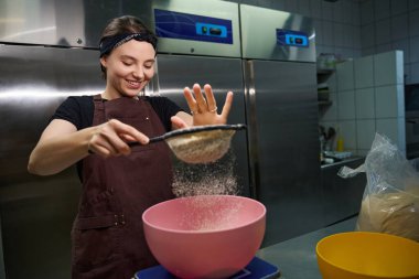 Önlük giymiş gülümseyen kadın fırıncı mutfak terazisine konmuş elek içinden badem unu seçiyor.