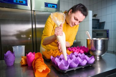 Baker, mutfak tezgahına yerleştirilmiş kek tepsisiyle boru torbasından kek kalıplarına hamur sıkıyor.