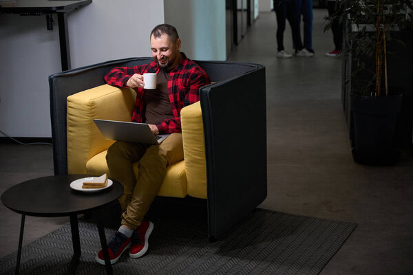 Позитивный кавказский мужчина пьет кофе и работает на ноутбуке в удобном коворкинге с Wi-Fi, готовит вкусный сэндвич, чтобы перекусить во время работы