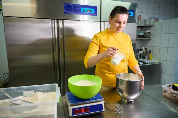 Focused pastry cook in uniform pouring milk from measuring cup into bowl