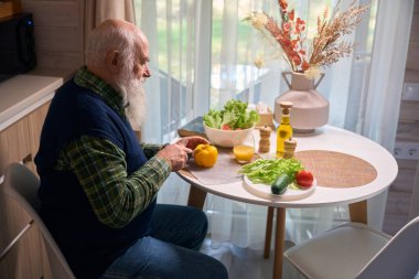 Mutfak masasındaki gri sakallı adam salata için sebze kesiyor, sıcak mavi bir yelek giyiyor.