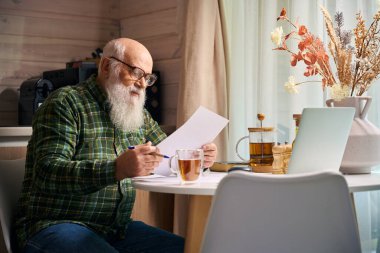 Büyükbabam mutfak masasında dizüstü bilgisayar ve belgelerle oturuyordu. Masada kurumuş çiçek kompozisyonu vardı.
