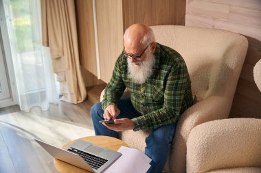 Gri saçlı yaşlı adam oturma odasında belgelerle çalışıyor. Dizüstü bilgisayar ve cep telefonu kullanıyor.