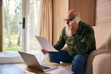 Gri sakallı yaşlı adam oturma odasında belgelerle çalışıyor. Dizüstü bilgisayar ve cep telefonu kullanıyor.