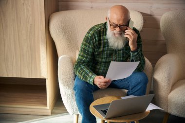 Yaşlı adam oturma odasında belgelerle çalışıyor, modern aletler kullanıyor.