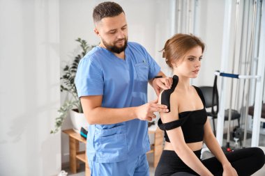 Kinesiyoterapi kliniğindeki doktor bandı hastaların omuzlarına uygular. Mavi üniformalı bir adam.